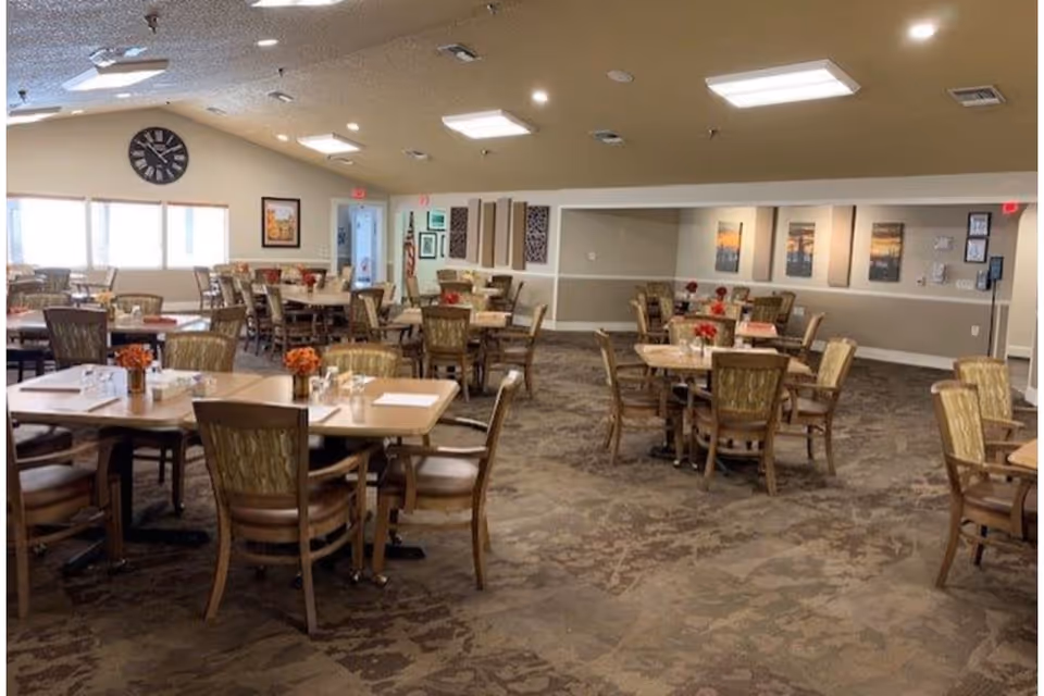 A spacious dining room with multiple wooden tables and chairs arranged neatly. Each table has a small floral centerpiece and place settings with napkins and glasses. The room has a carpeted floor, beige walls with framed artwork, and a large clock on one wall. Several windows allow natural light to enter the room.