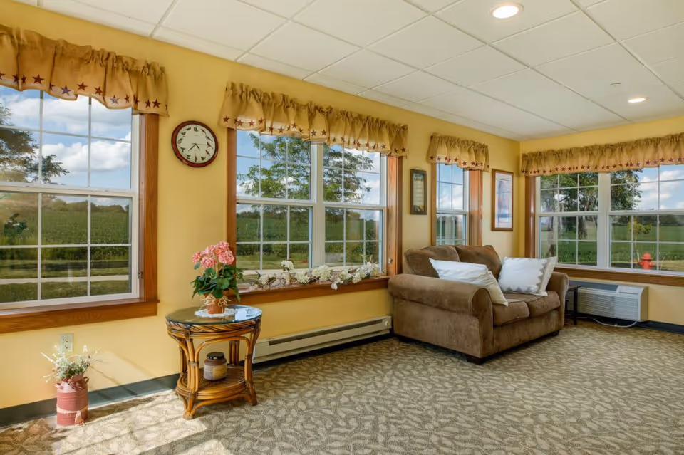 A cozy living room area with large windows showing a green outdoor landscape. The room has yellow walls, a brown loveseat with white pillows, a small round wooden table with a pink flower pot, and beige patterned carpet. The windows have yellow valances with red stars, and there is a clock on the wall between two windows.
