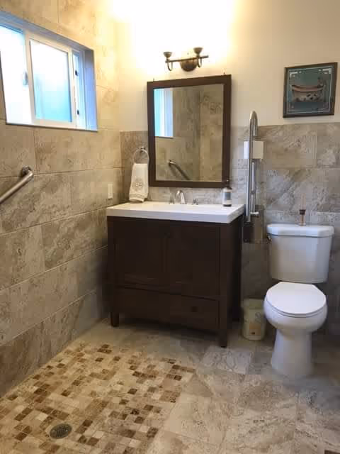 A bathroom with beige tiled walls and floor, featuring a walk-in shower area with a drain, a dark wooden vanity with a white sink and a mirror above it, a toilet with a grab bar beside it, a small window, and a wall-mounted light fixture above the mirror.
