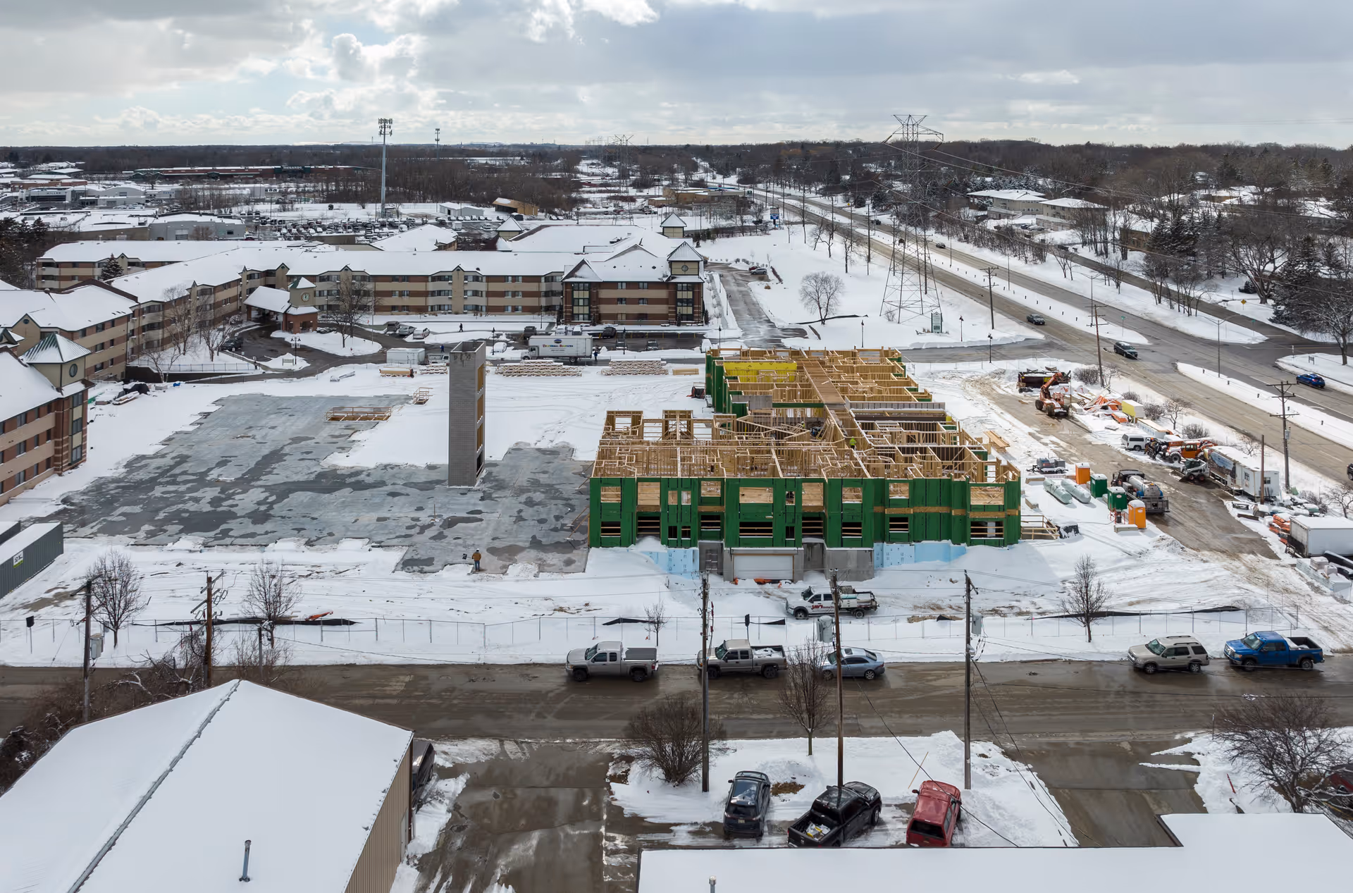 Aerial view of a senior living community named Forest Ridge Senior Community during winter. The image shows a large building complex with snow-covered roofs and surrounding ground. Part of the complex is under construction with wooden framing visible. Several vehicles are parked along the street in front of the buildings, and a road runs parallel to the community. Snow blankets the entire area under a cloudy sky.