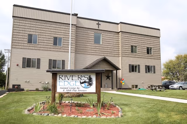 Exterior view of a three-story beige assisted living facility building with multiple windows and a small entrance porch. In front of the building is a landscaped area with a wooden sign that reads 'Rivers Edge Assisted Living Inc. & Adult Day Services.' There are a few balloons near the entrance and a couple of parked cars on the right side.