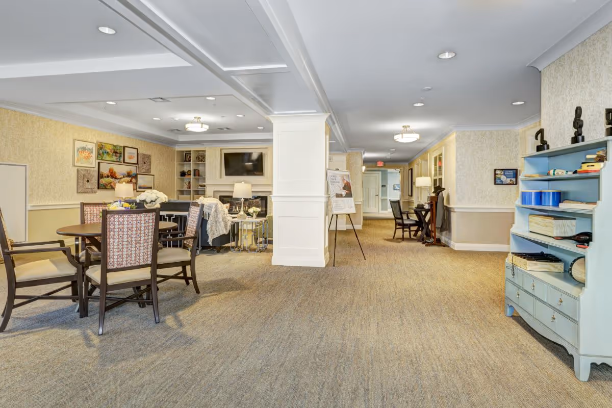 Bright senior living common area with round dining tables and chairs, lounge seating by a TV and fireplace, and shelving along the walls.
