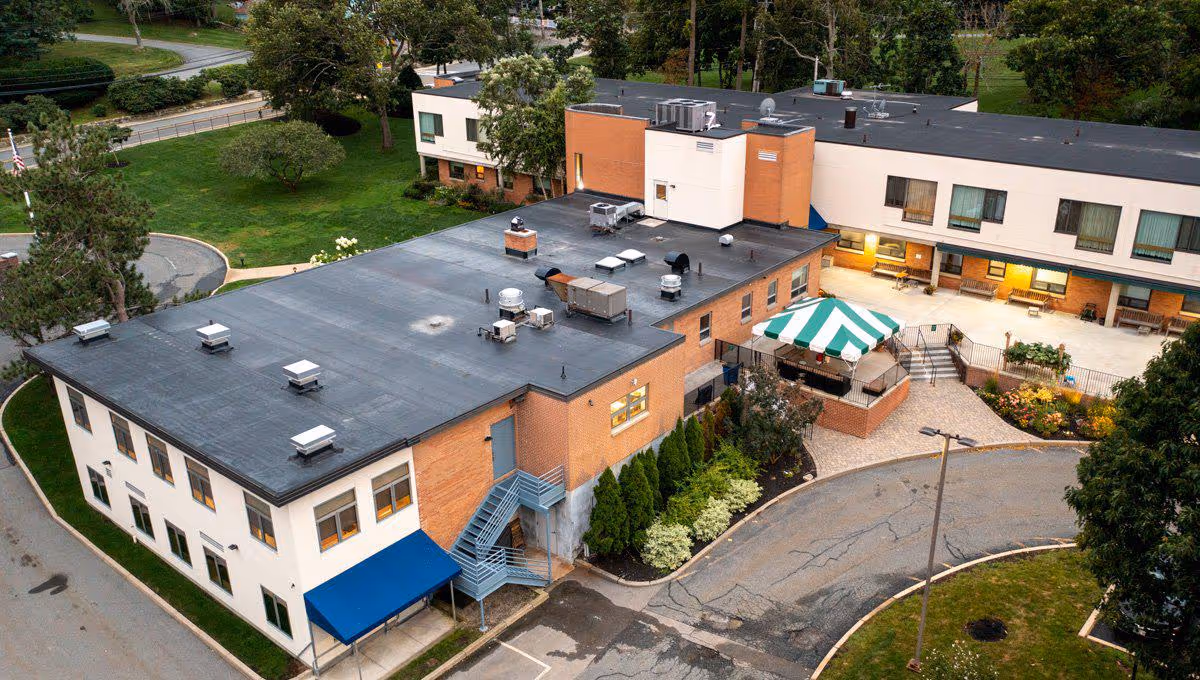 Aerial view of the Armenian Nursing & Rehabilitation Center showing a two-story building with a flat roof, surrounded by greenery and a curved driveway. The building has a brick and white exterior with several windows, a blue awning, and a green and white striped canopy over an outdoor patio area.