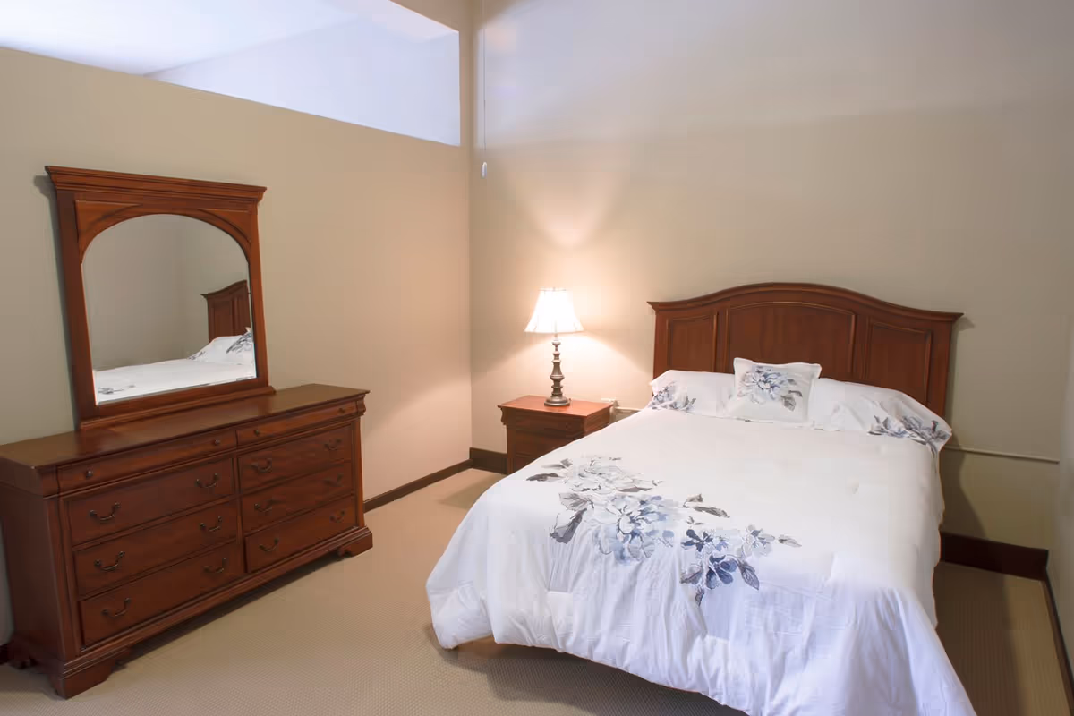 A bedroom with a wooden bed frame and a white comforter featuring a blue and gray floral pattern. There is a matching wooden nightstand with a lit lamp on it to the right of the bed. On the left side of the room, there is a wooden dresser with multiple drawers and a large mirror attached. The walls are painted beige and the floor is carpeted.