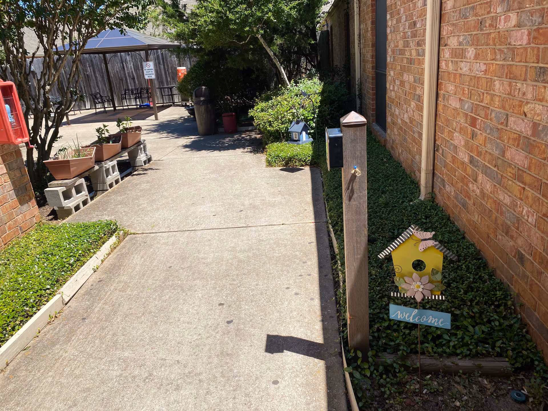 Outdoor walkway beside a brick building with green ground cover and small decorative birdhouses, one with a 'welcome' sign. There are potted plants on cinder blocks to the left, a trash can, and a shaded seating area with chairs and a table in the background.
