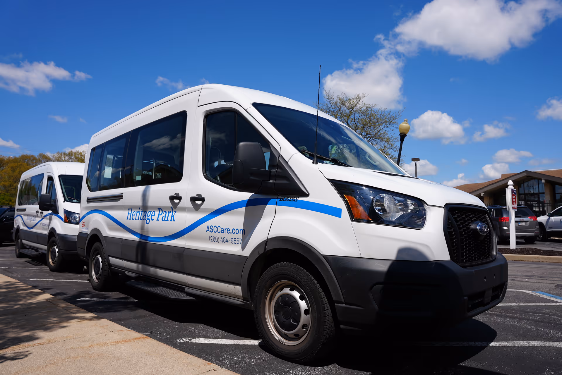 Two white shuttle vans parked in a parking lot under a blue sky with some clouds. The vans have blue wave graphics and the text 'Heritage Park' and 'ASCCare.com (260) 484-9557' on the side. A building and some cars are visible in the background.