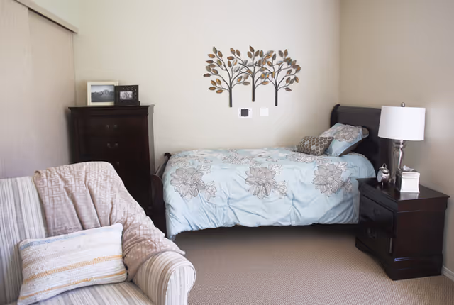 A cozy bedroom featuring a single bed with floral bedding, a dark wooden nightstand with a lamp and tissue box, a matching tall dresser with framed photos on top, a comfortable striped armchair with a throw blanket and pillow, and a decorative metal wall art of three trees above the bed.