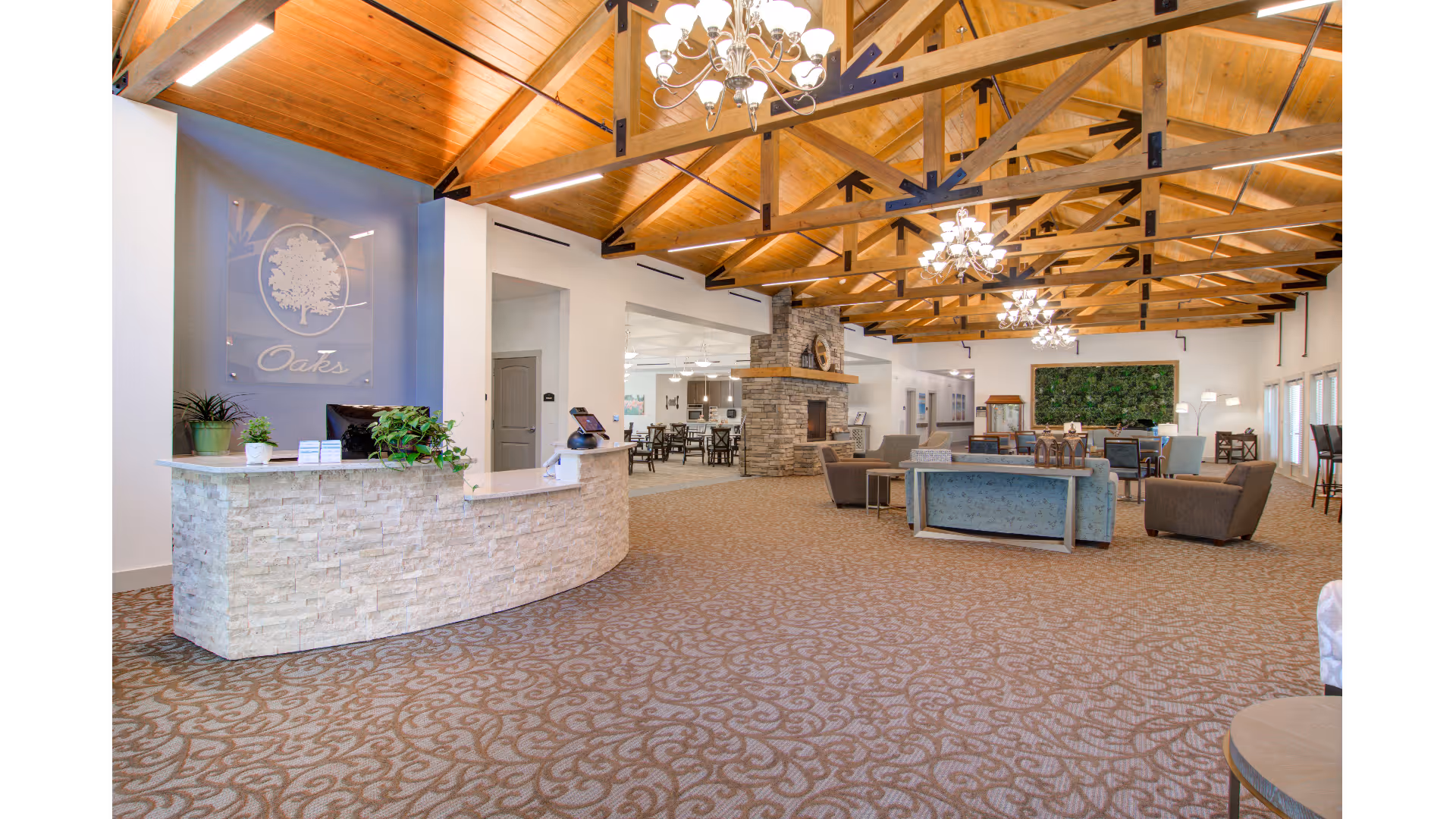 Spacious senior living facility common area with a stone reception desk on the left, a high wooden ceiling with exposed beams and chandeliers, a stone fireplace in the center, and multiple seating areas with chairs and sofas. The room has patterned carpet flooring and large windows on the right side.