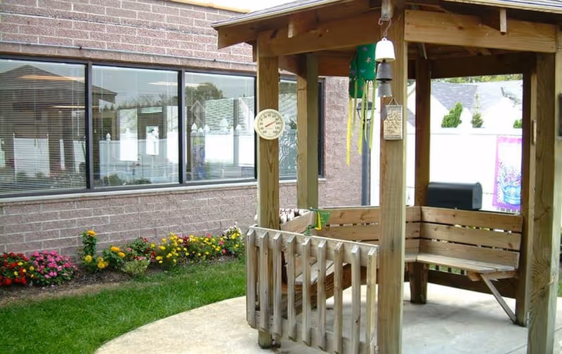 Wooden gazebo with a built-in bench, hanging thermometer and wind chimes in a small courtyard garden next to a brick building and flower beds.