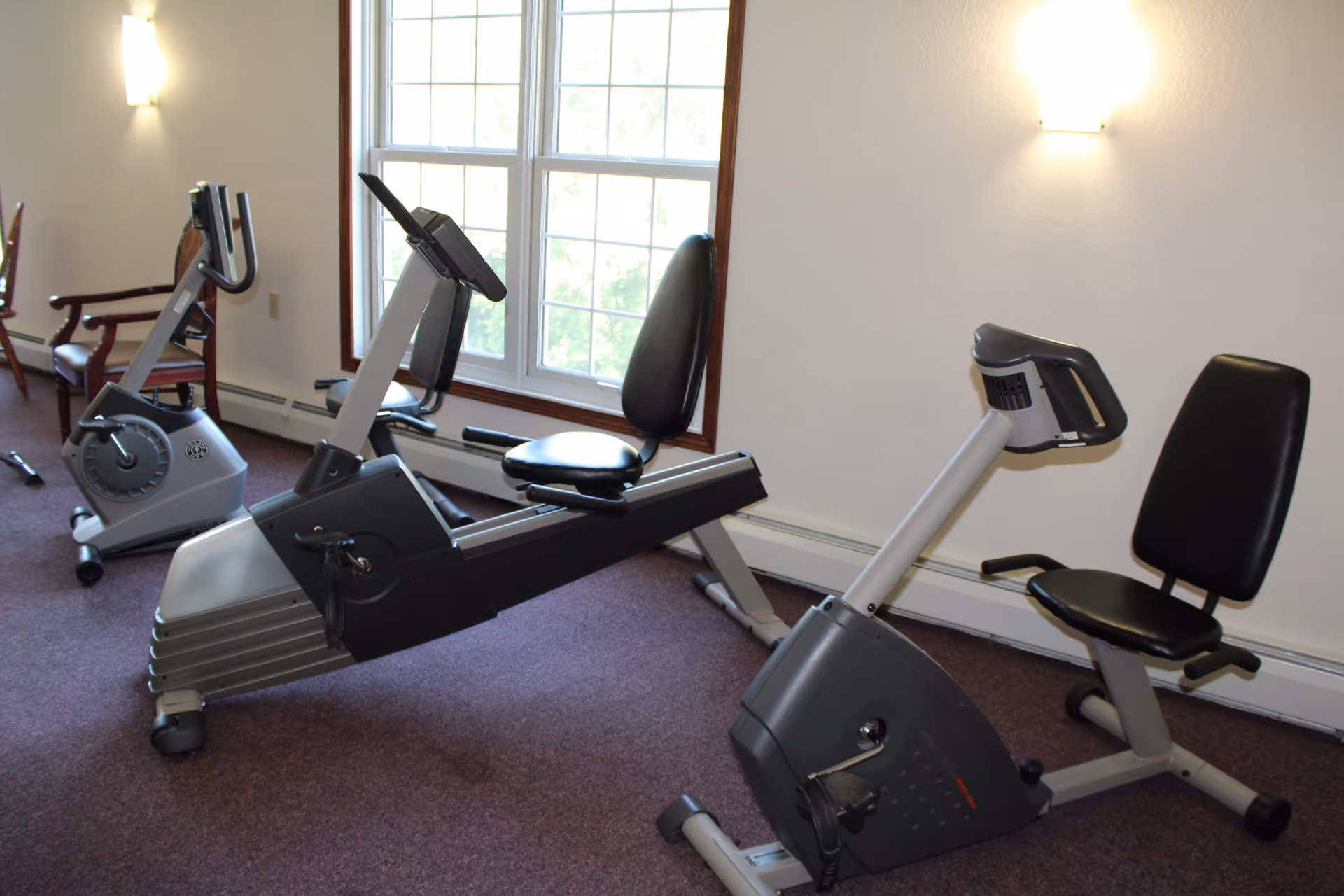 Carpeted exercise room with three recumbent stationary bikes positioned near a large window and wall sconces.