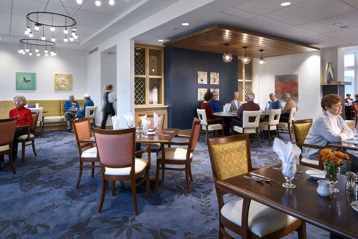 A dining room in a senior living facility with several elderly people seated at tables, some engaged in conversation. The room features modern lighting fixtures, artwork on the walls, and a mix of upholstered chairs and banquette seating. Tables are set with napkins, glasses, and silverware, and a staff member is walking through the space.
