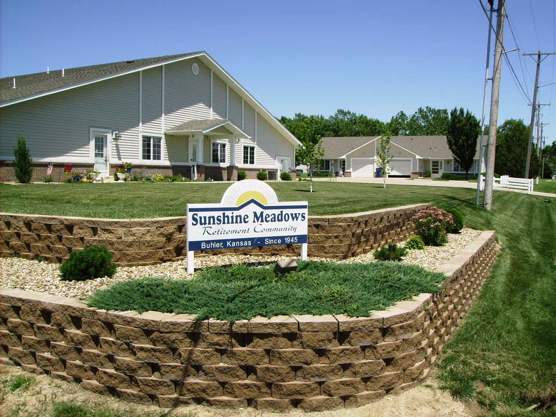 Exterior view of Sunshine Meadows Retirement Community showing a landscaped area with a stone retaining wall, green grass, shrubs, and a sign displaying the community name and location. Two light-colored buildings with pitched roofs are visible in the background under a clear blue sky.