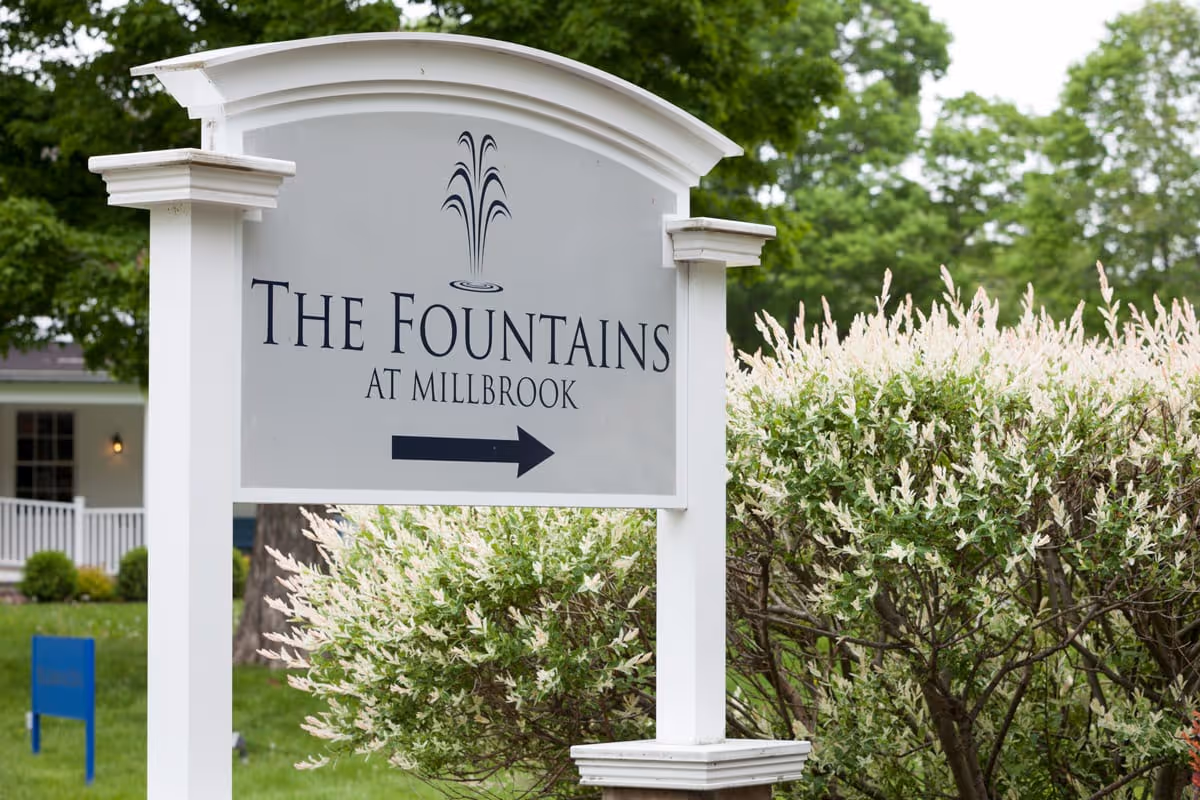 A white and gray sign with a fountain graphic and the text 'The Fountains at Millbrook' with an arrow pointing to the right, surrounded by green bushes and trees in an outdoor setting.