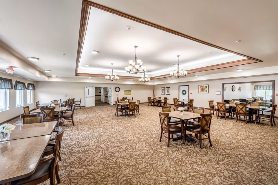 A spacious dining room with multiple wooden tables and chairs arranged neatly on a patterned carpet. The room features large windows with valances on the left side, several chandeliers hanging from a recessed ceiling with wood trim, and framed artwork on the walls. There is a doorway at the back leading to another room and an American flag near the far wall.