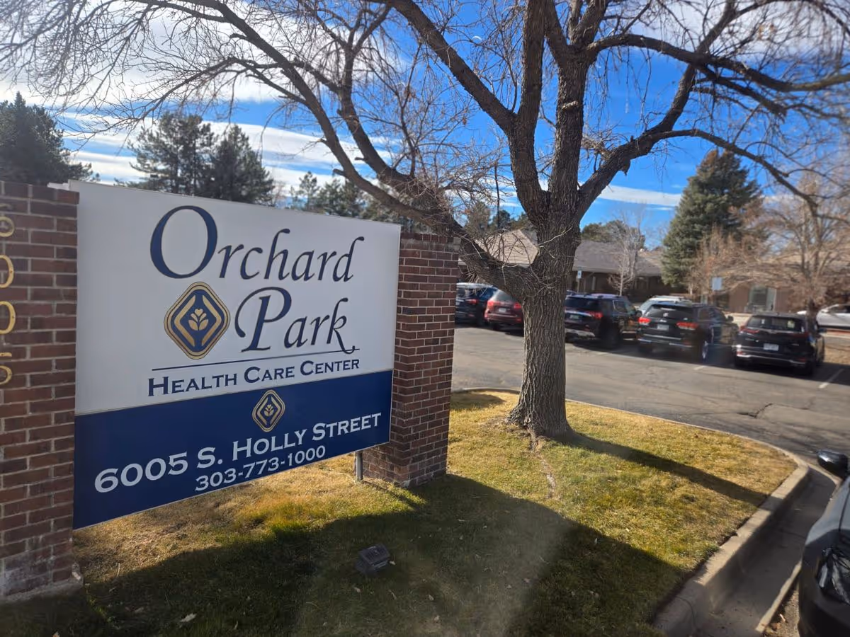 Outdoor sign reading 'Orchard Park Health Care Center' by a brick pillar with a tree, parking lot, and facility building in the background.