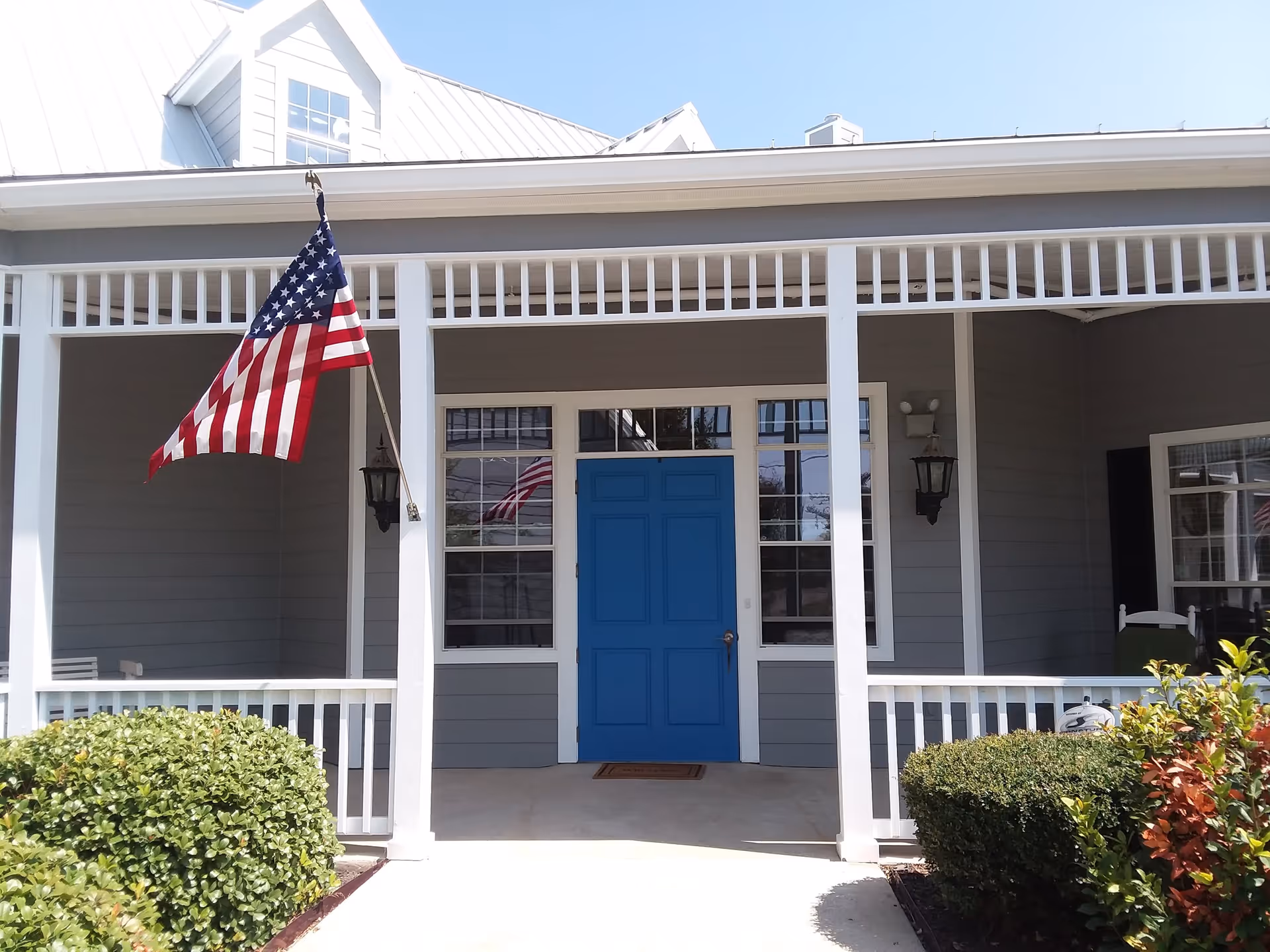 Front porch of a building with a bright blue door, two windows on either side, an American flag hanging on the left, white railings, and neatly trimmed bushes in front.
