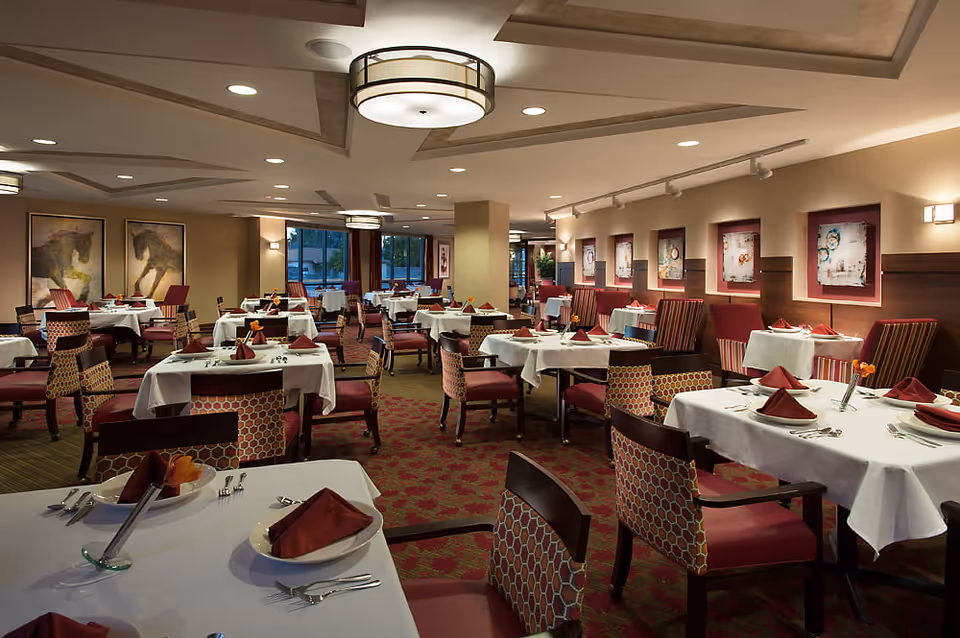 A spacious dining room with multiple tables covered in white tablecloths, each set with maroon napkins, plates, and silverware. The room features patterned chairs and booth seating along the wall, with framed artwork hanging above. The ceiling has recessed lighting and modern circular light fixtures, and large windows allow natural light to enter.