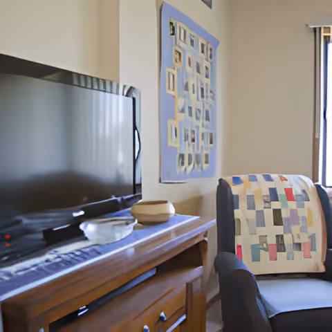 A cozy living room corner featuring a flat-screen TV on a wooden cabinet with decorative items, a cushioned armchair draped with a colorful patchwork quilt, and a wall hanging with a geometric pattern. Natural light comes through a window with vertical blinds.
