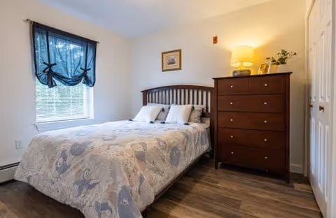 A cozy bedroom with a bed covered in a patterned quilt, two pillows, a wooden headboard, a wooden dresser with a lamp and small decorative items on top, a window with a dark curtain, and a framed picture on the wall.