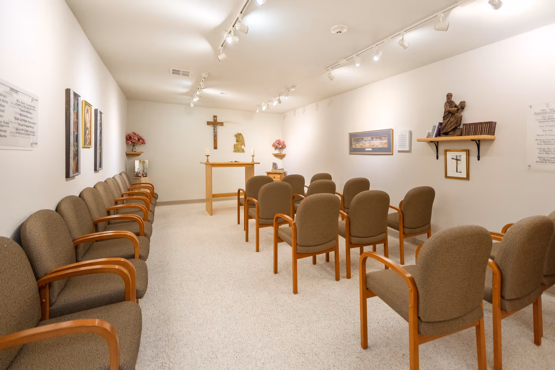 A small chapel room with rows of brown cushioned chairs with wooden armrests arranged facing a wooden altar. The altar has two candles and religious decorations including a crucifix on the wall behind it. The walls are white and decorated with religious artwork and plaques. The room is well-lit with ceiling track lighting.