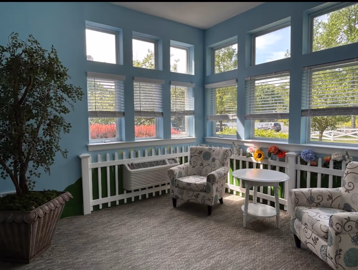 A bright sitting area with large windows letting in natural light. The walls are painted light blue with a white picket fence-style railing beneath the windows. Two patterned armchairs and a small white round table are arranged on a carpeted floor. A large potted plant is placed in the corner, and colorful artificial flowers decorate the railing.