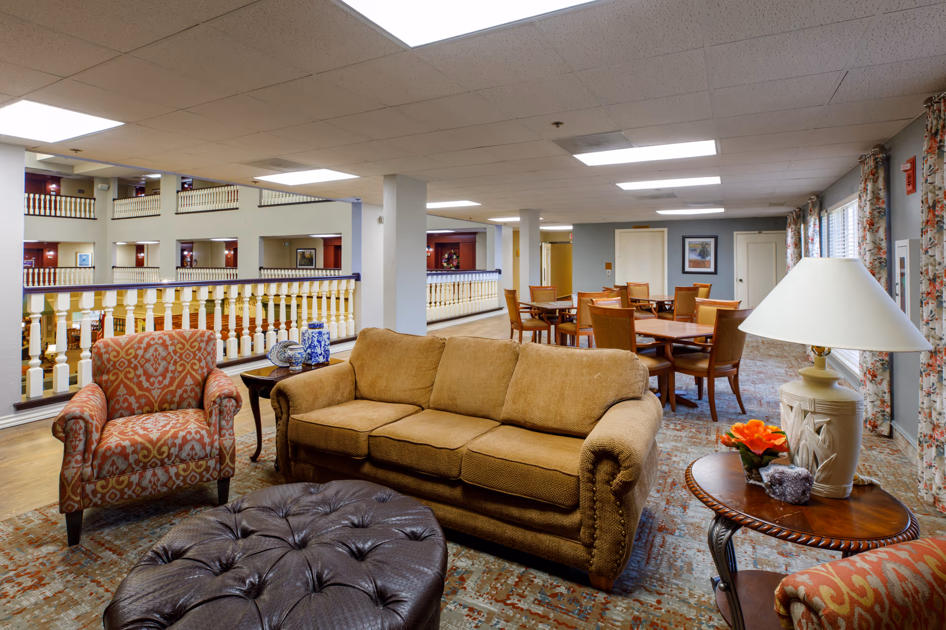 A spacious and well-lit common area in Rivermont Independent Living featuring a brown upholstered sofa, a patterned armchair, a large tufted leather ottoman, and a wooden side table with a lamp and a small flower arrangement. In the background, there are multiple wooden tables and chairs arranged for dining or social activities, with large windows covered by floral curtains letting in natural light.