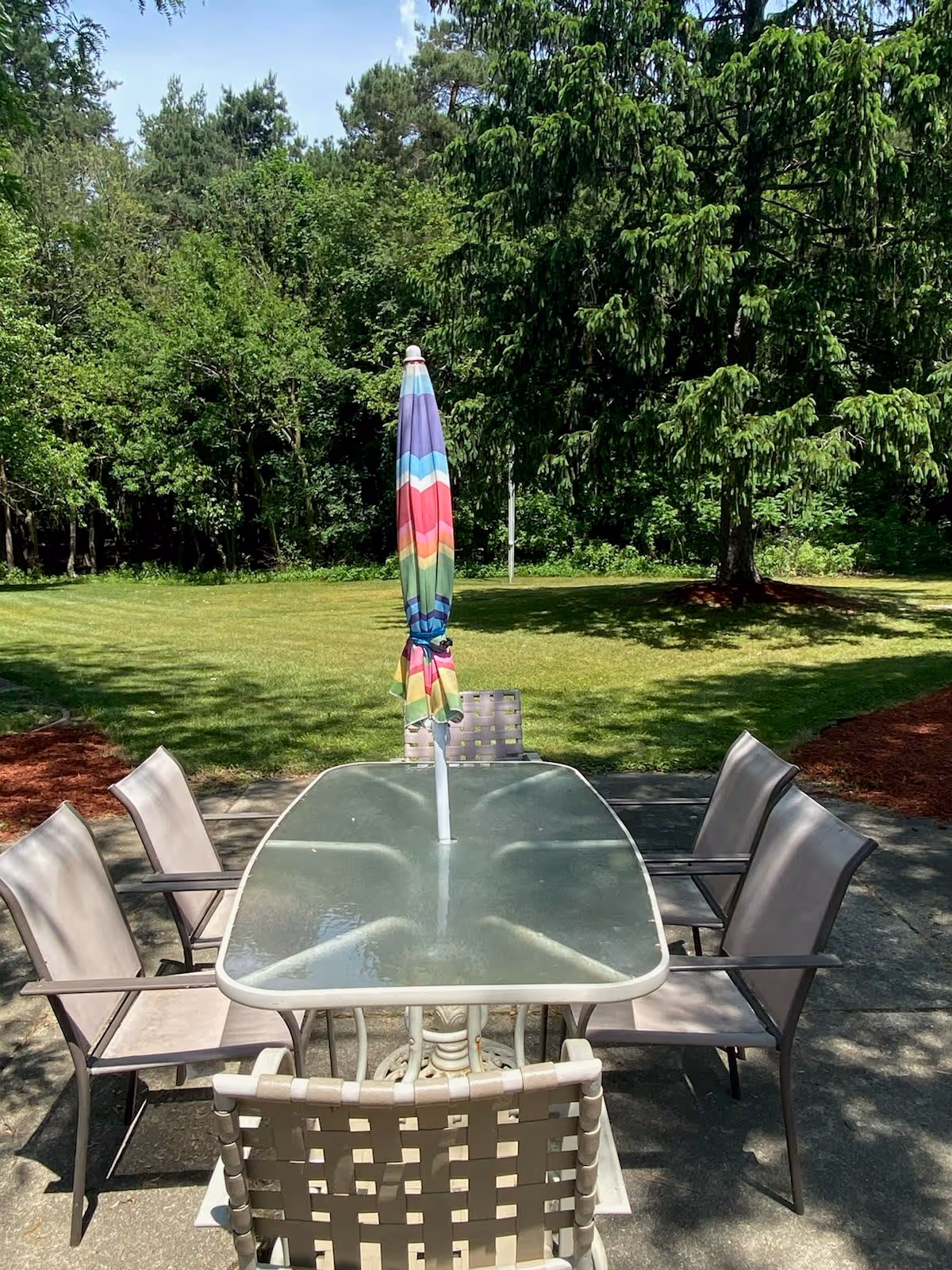 Outdoor patio area with a glass-top table surrounded by six beige chairs. A closed, colorful striped umbrella is positioned in the center of the table. The patio is surrounded by green grass and trees under a sunny sky.
