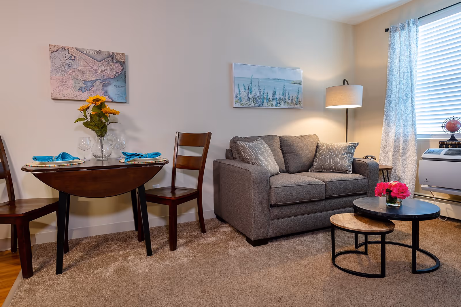 A cozy living area featuring a small wooden dining table set for two with blue napkins and wine glasses, a gray upholstered loveseat with patterned pillows, a floor lamp, two nested round coffee tables with a small vase of pink flowers, and a window with white blinds and patterned curtains. Wall art includes a map of Boston and a coastal painting.