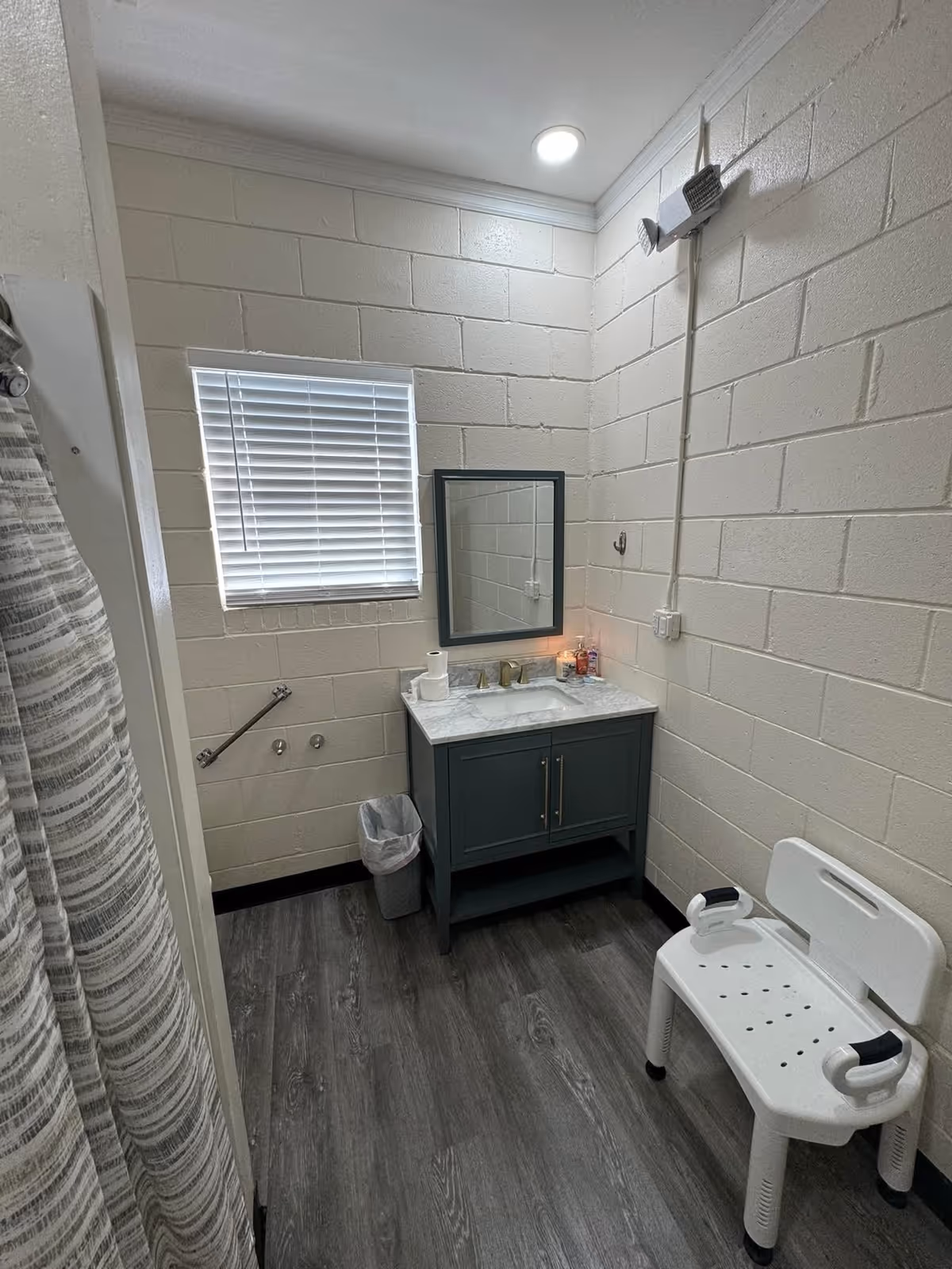 A small bathroom with white painted cinder block walls and gray wood-look flooring. There is a window with white blinds above a gray vanity with a marble countertop, a mirror, and a few items on the counter. A white plastic shower chair with armrests is positioned near the vanity. A shower curtain with gray and white stripes is partially visible on the left side. A grab bar is mounted on the wall near the window, and a small trash can is placed next to the vanity.