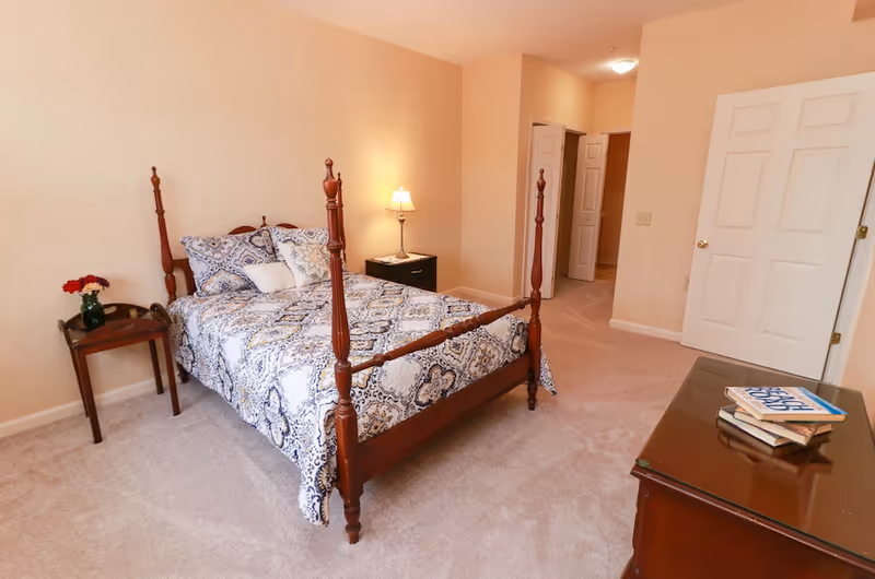 A bedroom with a wooden four-poster bed covered with a patterned bedspread and pillows. There is a small wooden side table with a vase of flowers on the left and a nightstand with a lamp on the right. The room has beige walls and carpeted floor, with two white doors visible in the background and a wooden dresser with books on top in the foreground.