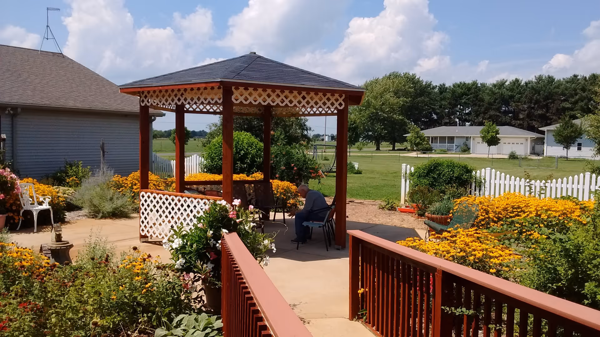 A peaceful outdoor garden area with a wooden gazebo featuring white lattice trim. There are colorful flowers and green shrubs surrounding the paved walkway and seating area. An elderly person is sitting on a chair under the gazebo. In the background, there are houses, trees, and a clear blue sky with some clouds.