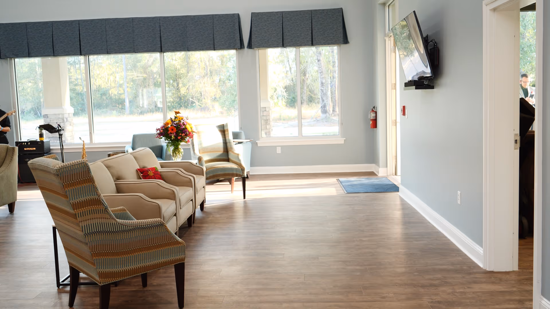 A bright and spacious living room area with large windows letting in natural light. The room features a beige sofa, two patterned armchairs, a bouquet of flowers on a small table, and a wall-mounted TV. The floor is wooden, and the walls are painted light blue. There is a fire extinguisher mounted on the wall near the entrance.