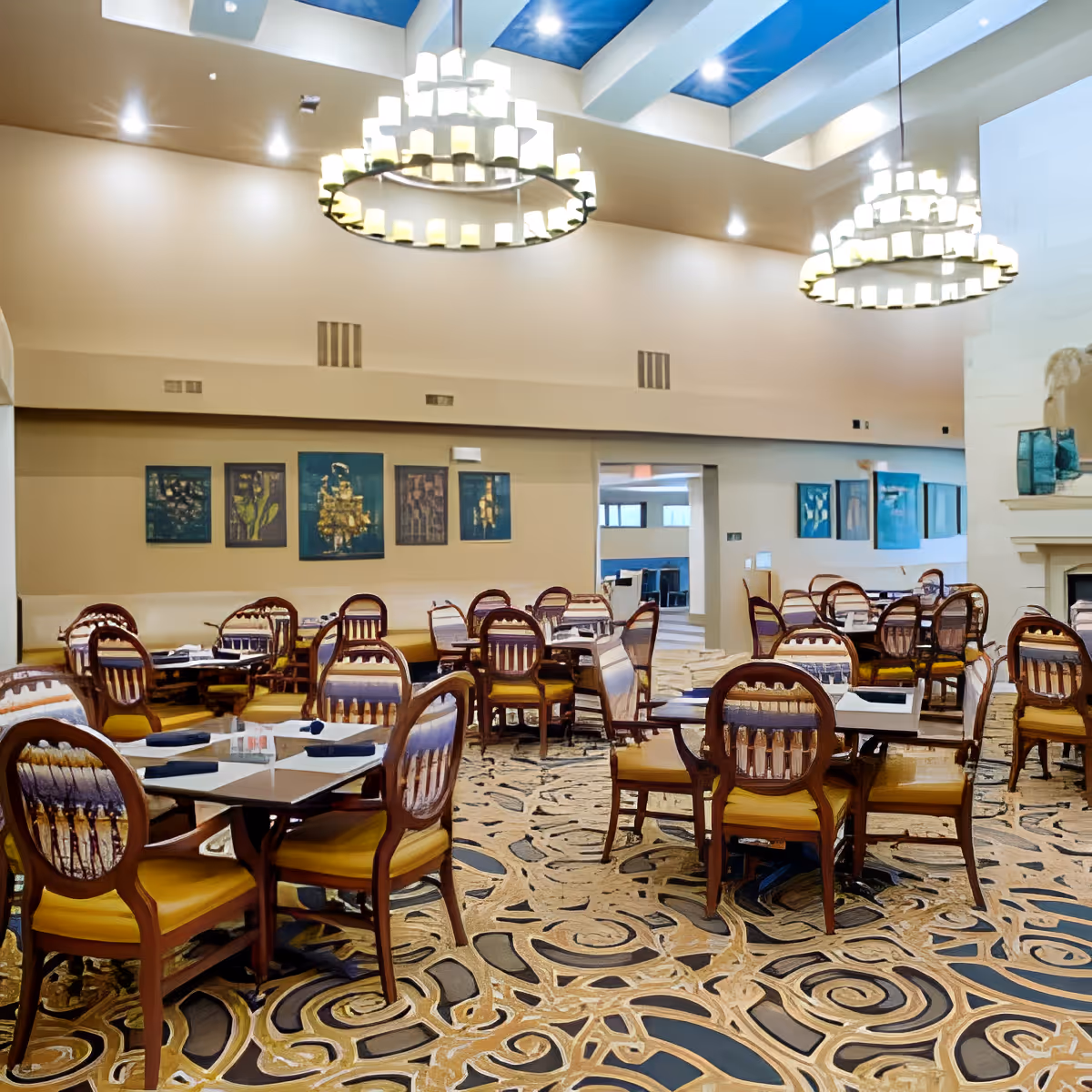 A spacious dining room with multiple wooden tables and chairs featuring yellow cushions. The room has a patterned carpet, beige walls adorned with framed artwork, and two large circular chandeliers hanging from a ceiling with blue accents. There is a fireplace on the right side and an open doorway leading to another area.