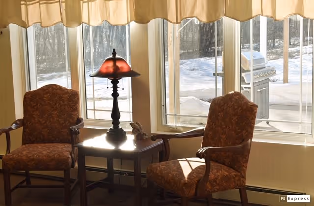 Two upholstered armchairs with wooden frames positioned on either side of a small wooden table with a decorative lamp and a small figurine, set in front of large windows showing a snowy outdoor scene with a barbecue grill.
