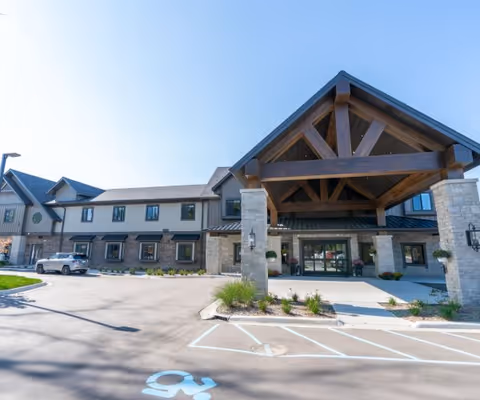 Exterior view of Grace Senior Living Rochester Hills building with a large covered entrance supported by stone pillars and wooden beams. The building has multiple windows and a parking area with a handicapped parking space in front.