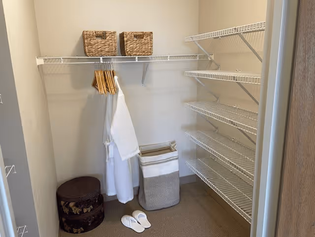 A walk-in closet with white wire shelving on the right wall and a hanging rod on the back wall holding wooden hangers and two white robes. On the floor, there are two round decorative storage boxes, a striped laundry basket, and a pair of white slippers. Two woven baskets are placed on the top shelf.