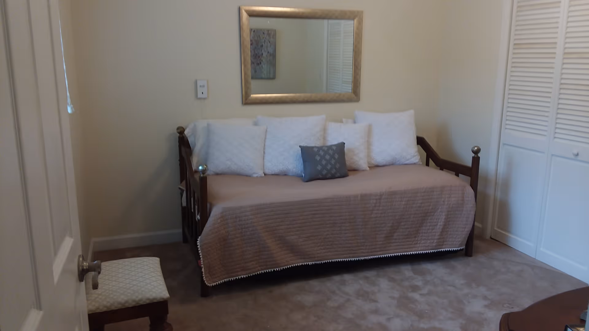 A small bedroom with a daybed covered in a beige quilt and five pillows, four white and one gray. Above the bed is a rectangular mirror with a gold frame. To the right is a white closet with louvered doors, and to the left is a partially open door with a small cushioned stool nearby. The room has beige walls and carpeted floor.