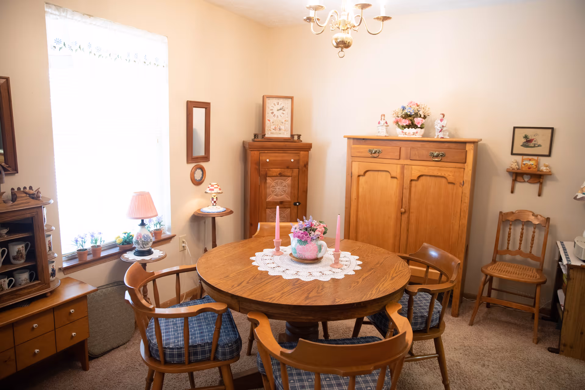 A cozy dining room with a round wooden table surrounded by four wooden chairs with blue plaid cushions. The table is decorated with a white lace doily, a floral centerpiece, and two pink candles. The room has beige walls and carpeted floor. There is a window with sheer curtains letting in natural light, a small side table with a lamp, a wooden cabinet, and various decorative items including framed pictures and small figurines.