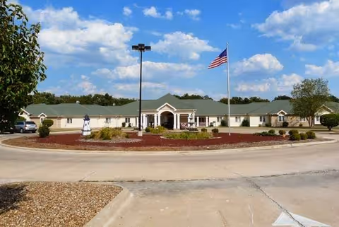 Single-story building with a green roof and beige exterior walls, surrounded by a circular driveway with landscaped mulch and bushes. An American flag is flying on a flagpole in front of the building under a partly cloudy blue sky.