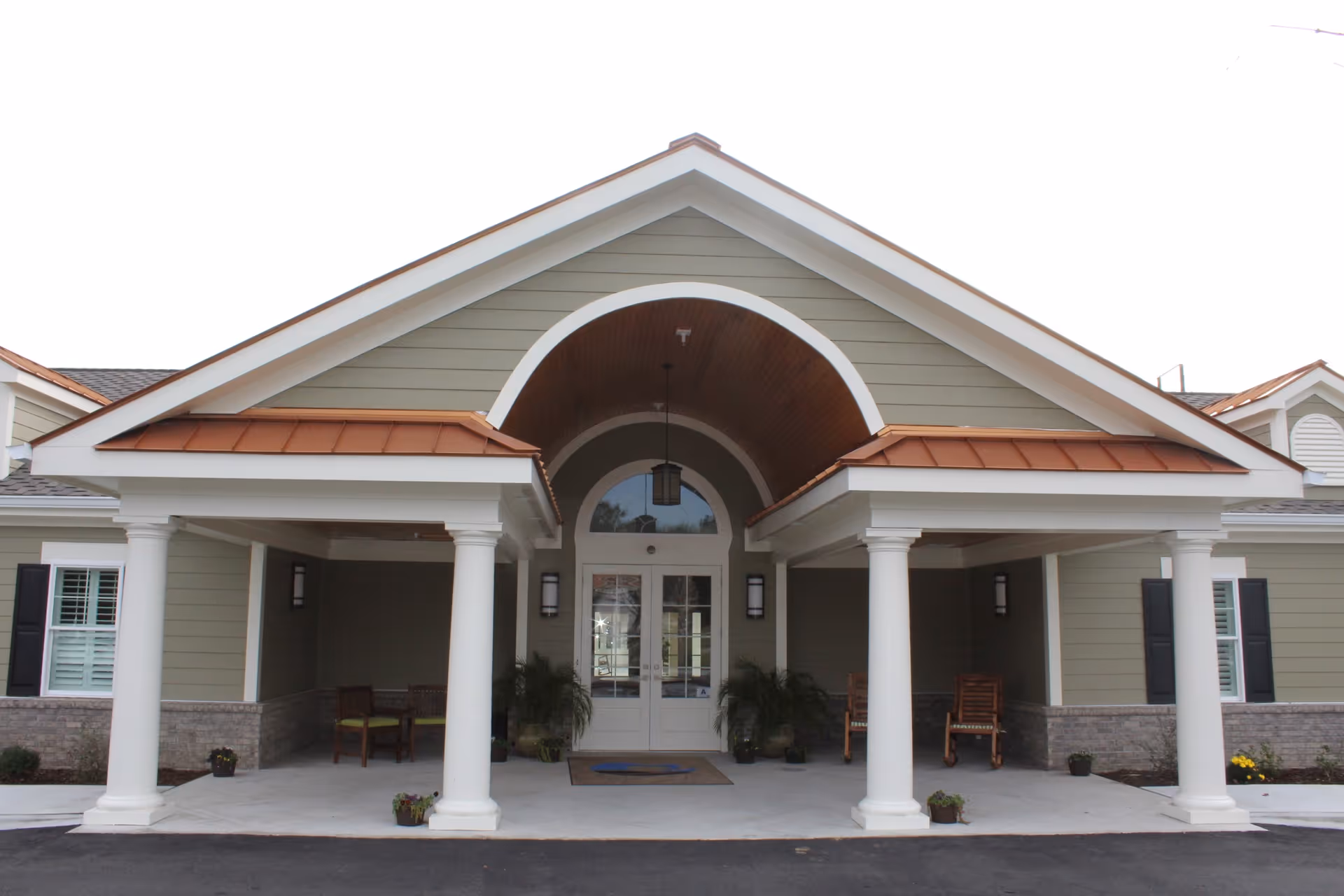 Front entrance of a senior living facility with a covered porch supported by white columns, green siding, and a copper-colored roof accent. There are chairs and potted plants on the porch area.