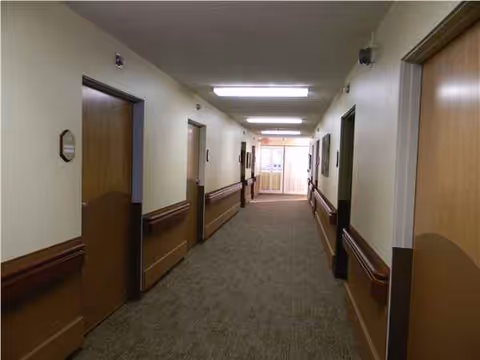 A long beige hallway in a senior living facility with doors, handrails, and carpet.