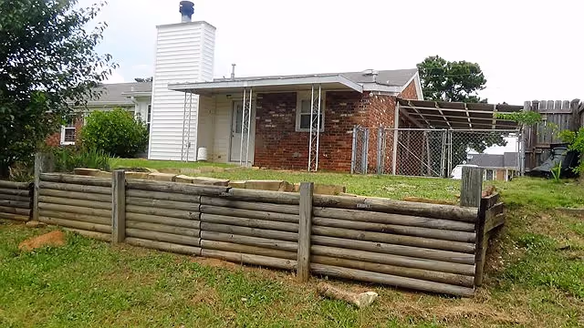 Exterior view of a single-story brick residential building with a white chimney and a small fenced area on the right side. The foreground shows a grassy area with a wooden retaining wall made of horizontal logs.