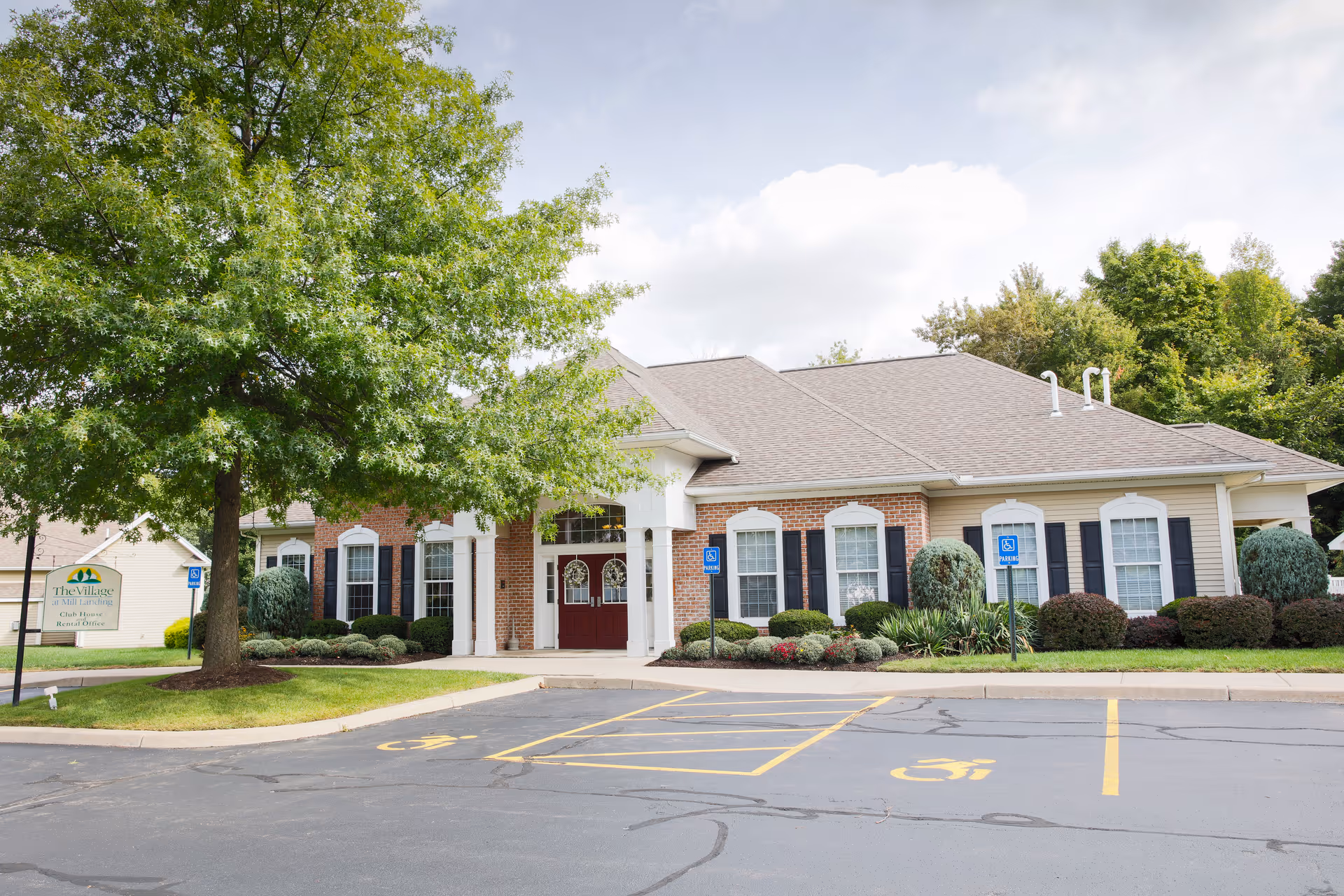 Exterior view of The Village Townhomes building with a brick and beige facade, multiple windows with black shutters, a red double door entrance, surrounded by landscaped bushes and trees, and a parking lot with marked handicap parking spaces in front.