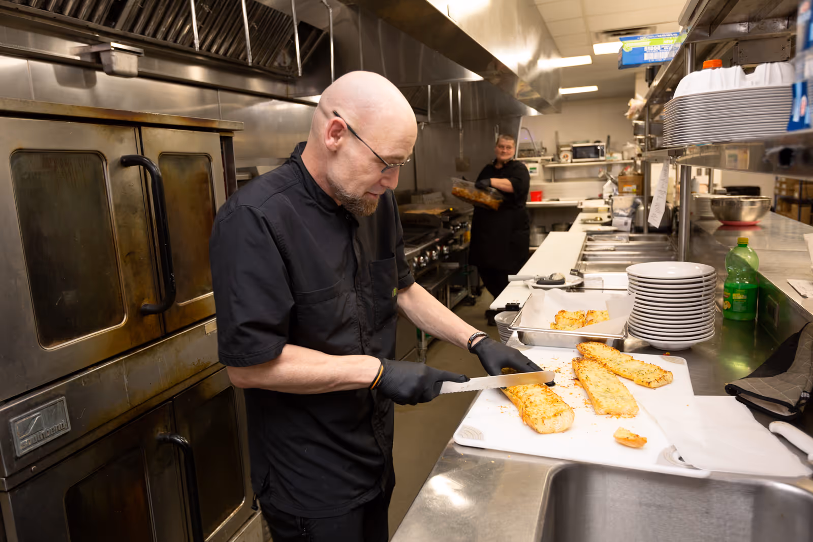 A man wearing black chef attire and gloves is slicing garlic bread on a white cutting board in a commercial kitchen. In the background, a woman also dressed in black chef attire is holding a tray and smiling. The kitchen has stainless steel appliances, countertops, and shelves with plates and kitchen supplies.