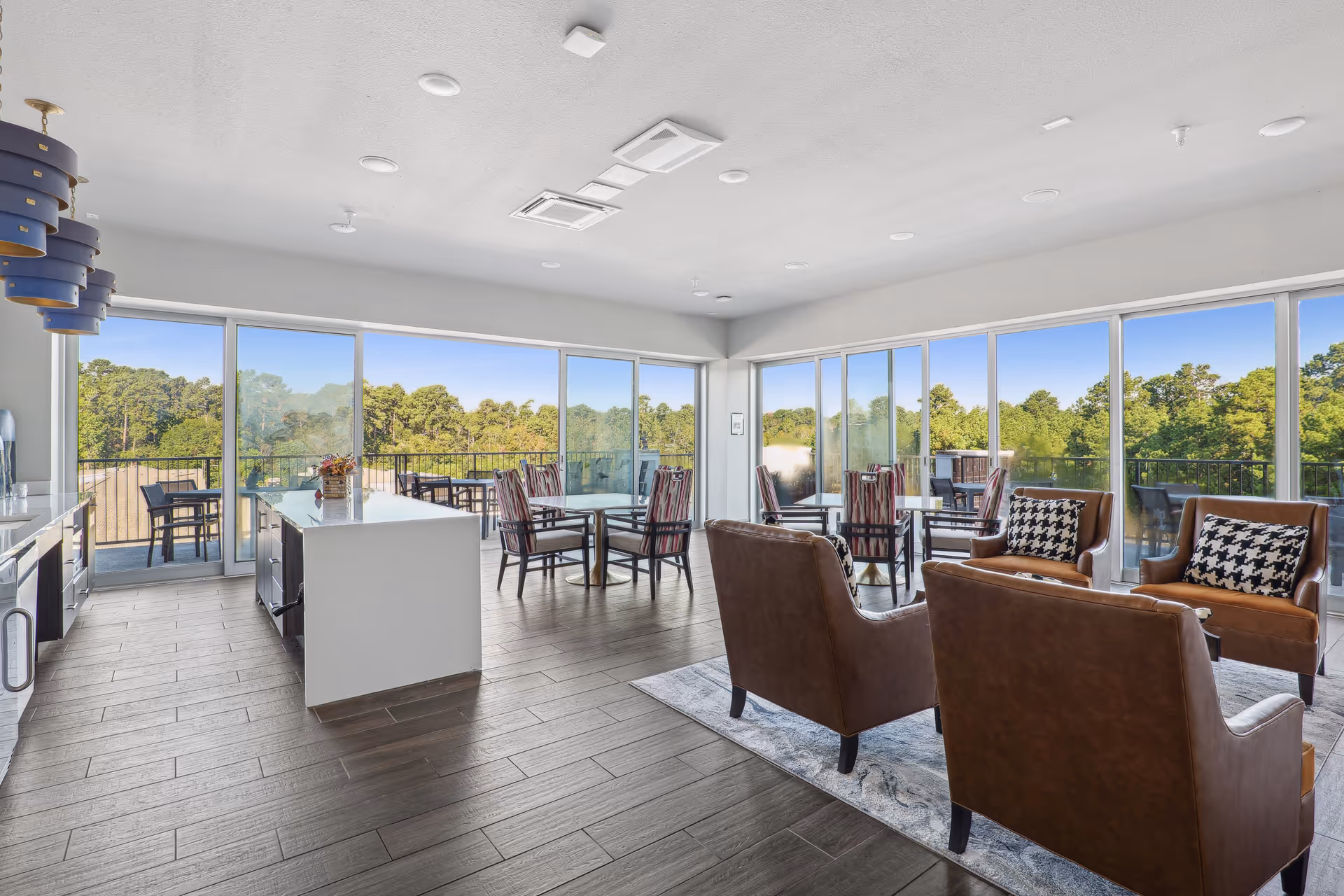 Bright communal lounge and dining area with a kitchen island, tables, leather chairs, and floor-to-ceiling windows overlooking trees.