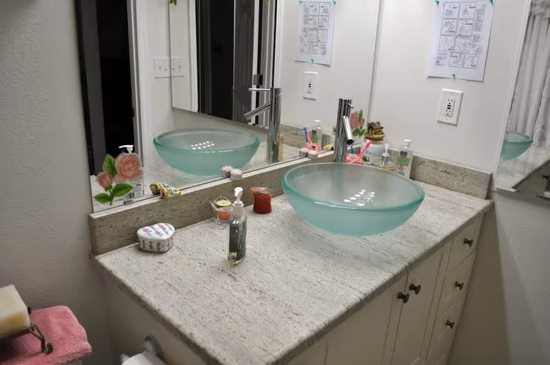 A bathroom countertop with a frosted glass vessel sink and a modern faucet. The countertop is light-colored granite with various items including a soap dispenser, a small container, a candle, and toothbrushes. A large mirror is mounted on the wall behind the sink, reflecting the items and part of the room. The walls are white, and there are two electrical outlets visible above the countertop.