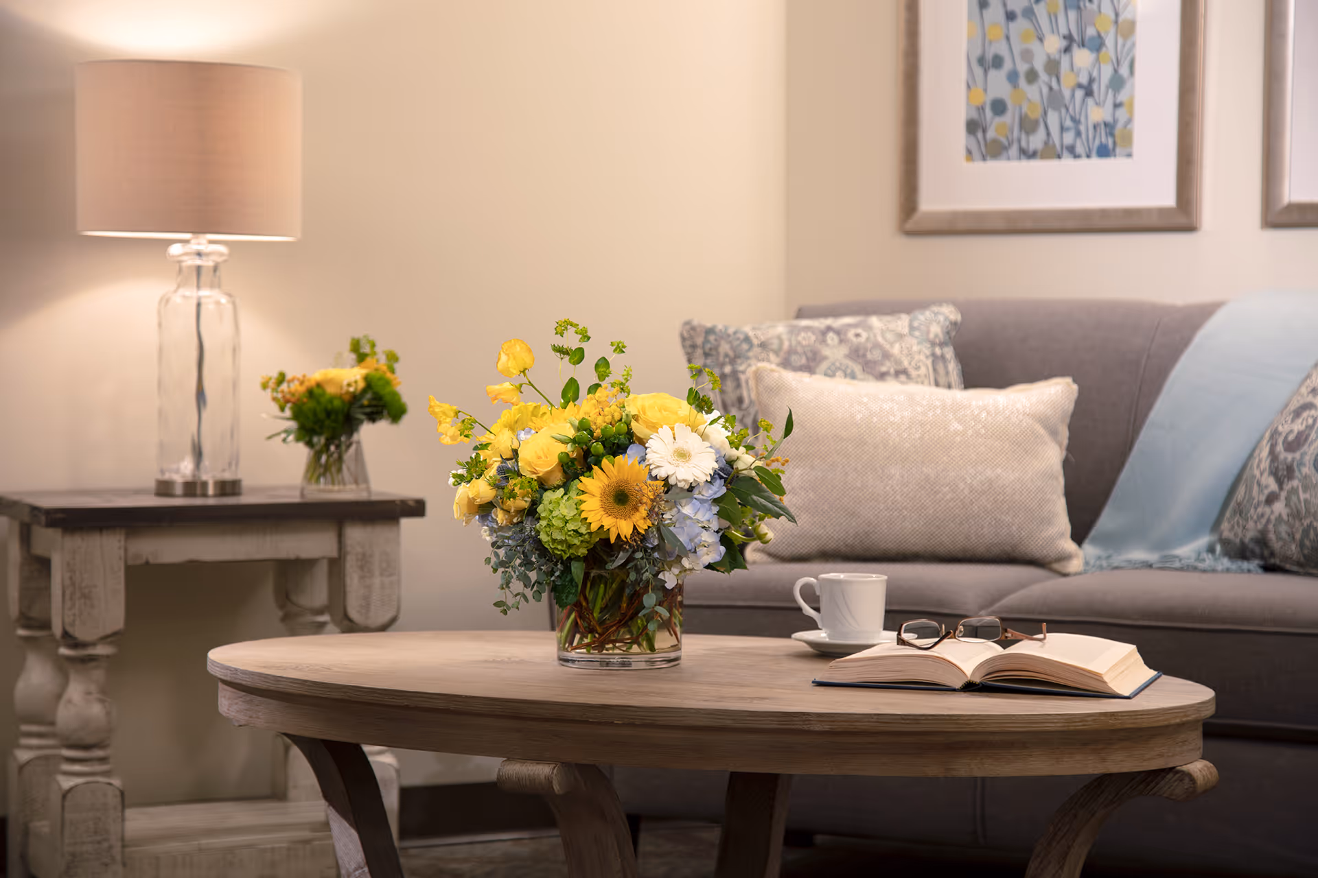 A cozy living room area with a gray sofa adorned with patterned and plain cushions, a light blue throw blanket, a wooden coffee table with a vase of yellow and white flowers, an open book with reading glasses, and a white coffee cup. In the background, there is a side table with a glass lamp and another small vase of flowers, and two framed floral artworks on the wall.