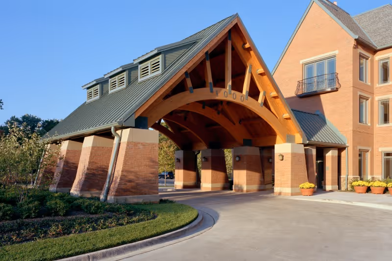 Entrance of a senior living facility with a large covered driveway supported by brick pillars and wooden beams, adjacent to a multi-story brick building with windows and flower pots near the entrance.