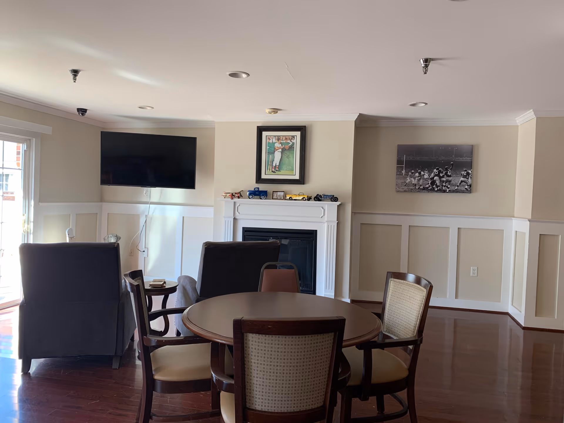 A cozy living room area with a round wooden table surrounded by four chairs in the foreground. Behind the table are two armchairs facing a white fireplace mantel decorated with small model cars and a framed picture above it. A flat-screen TV is mounted on the wall to the left, and a black and white sports photograph hangs on the wall to the right. The room has wooden flooring and beige walls with white wainscoting.