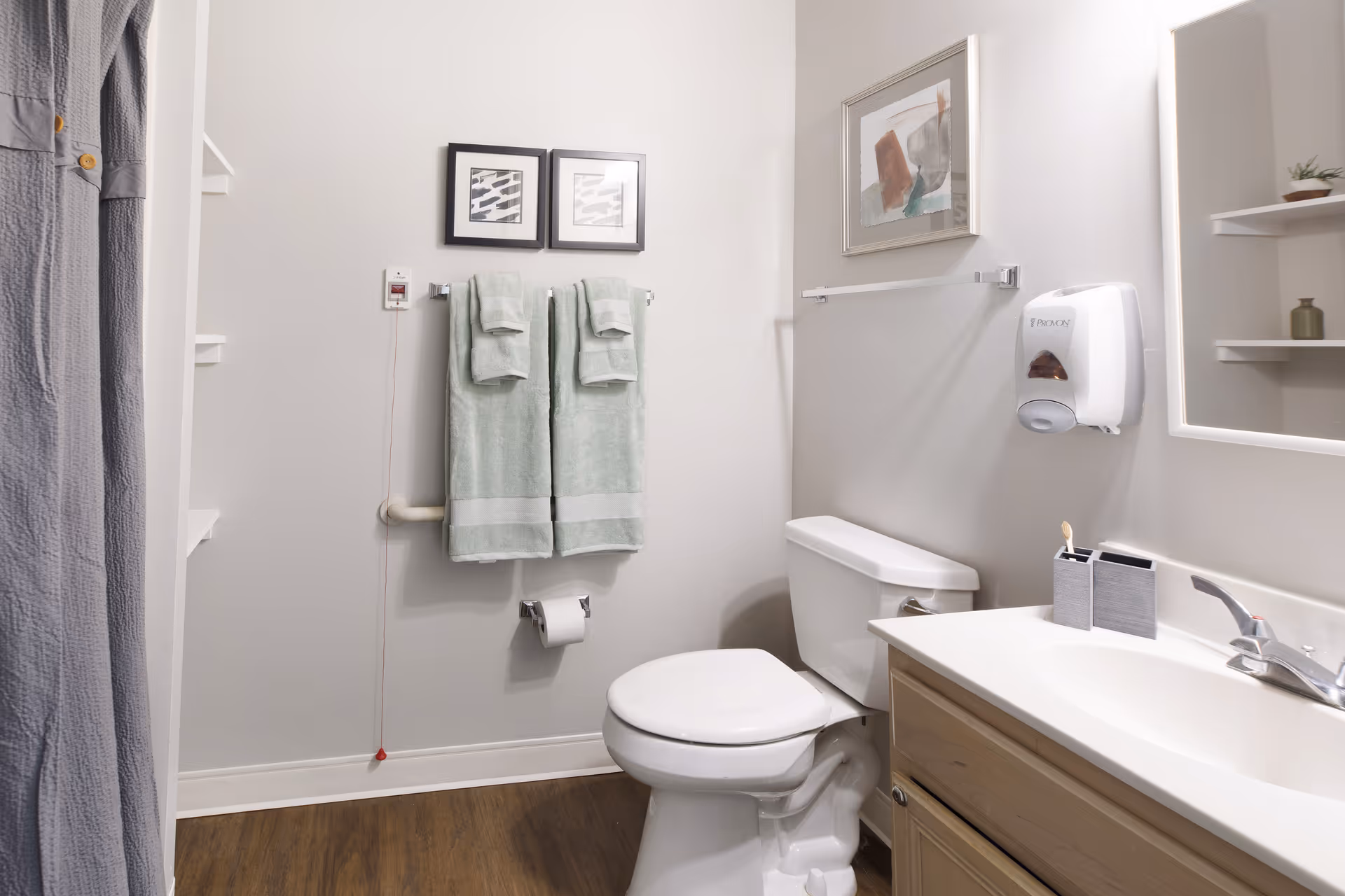 A clean and well-lit bathroom featuring a white toilet, a wooden vanity with a white sink, a wall-mounted soap dispenser, and a mirror with built-in lighting. Two green towels hang on a towel rack above the toilet, with two framed abstract artworks on the wall above the towels. A gray shower curtain is partially visible on the left side, and the floor has a wood-like finish.